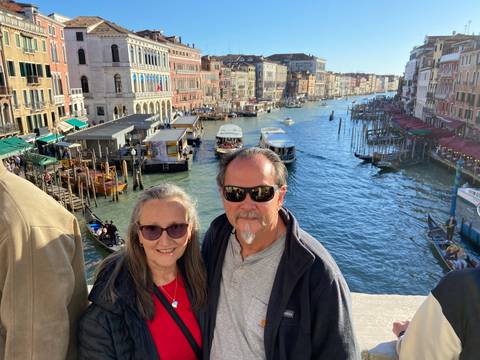       Couple leaning on a stone bridge railing with Venice’s Grand Canal bustling with boats behind.
  