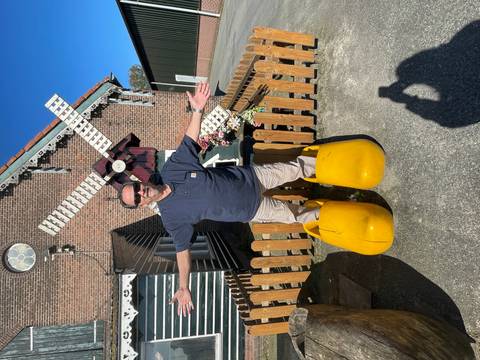       Man humorously wearing oversized yellow wooden clogs outside a small Dutch windmill workshop.
  