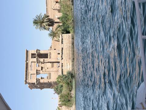       Ancient sandstone kiosk ruins on an island in the Nile viewed from a boat on bright blue water.
  