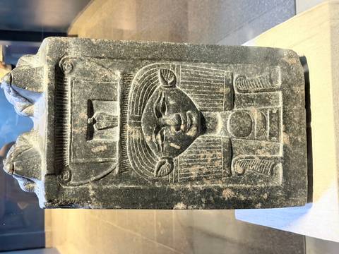       Stone Egyptian statue with carved female face displayed in a museum gallery.
  