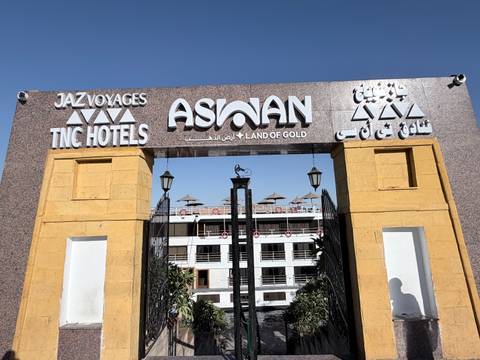       Entrance arch reading 'Aswan Land of Gold' leading to a docked Nile cruise ship under clear skies.
  