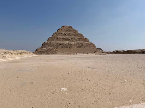       Wide view of the Step Pyramid of Djoser standing alone in a vast sandy courtyard under blue sky.
  