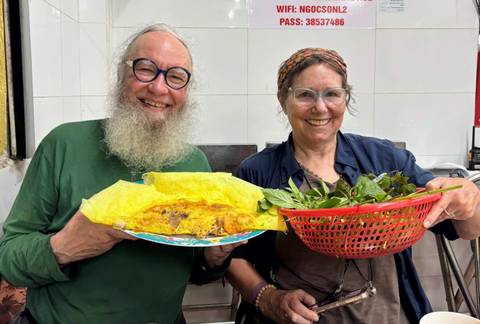      Smiling couple proudly displaying Vietnamese pancake and basket of herbs during a cooking class.
  