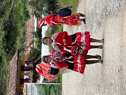       Two young girls in bright traditional Andean clothing walk along a rural road with villagers in the background
  