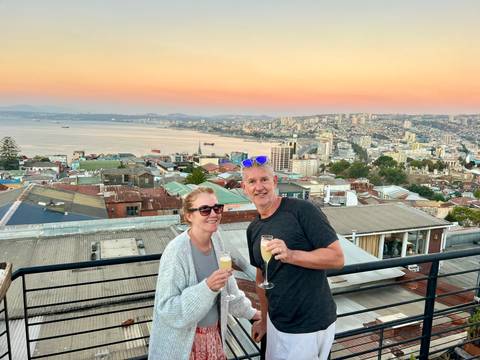       Smiling couple clinking champagne glasses on a rooftop terrace overlooking a coastal city at sunset
  