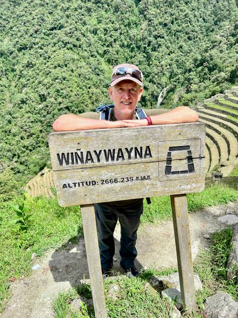       Traveler leaning on a weathered wooden sign reading 'Wiñaywayna' with terraced mountains behind
  