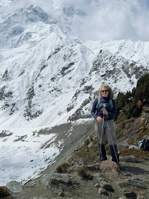       A smiling hiker poses with trekking poles against a backdrop of steep snow-covered slopes and conifers.
  