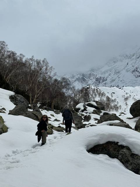       Two trekkers navigate boulders in a foggy, snow-covered alpine valley beneath looming cliffs.
  