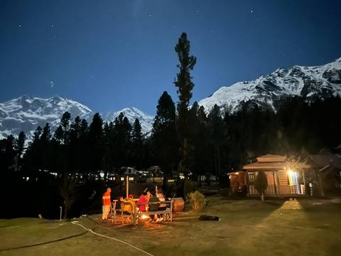       Under a starry moonlit sky, travellers sit around a campfire with snow-capped peaks towering above dark pines.
  