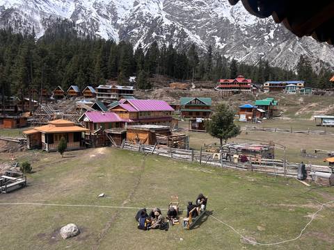       A rustic alpine settlement of colourful wooden cabins lies in a forested meadow beneath craggy snowy peaks.
  