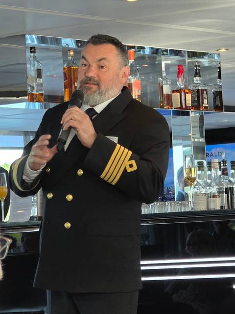       Ship captain in formal uniform speaking into a microphone inside a stylish cruise bar.
  