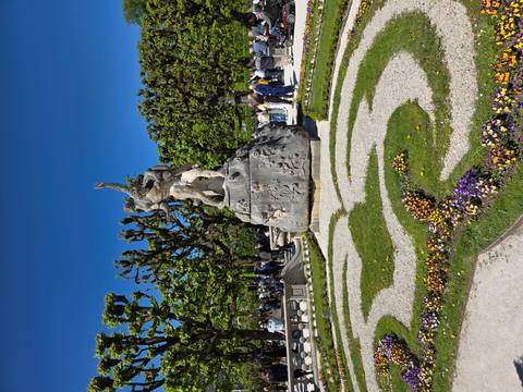       Stone fountain statue in manicured garden maze with crowds touring on a bright spring day.
  