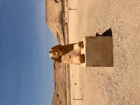       Small sphinx statue rests on a stone plinth in a vast sandy archaeological area under a blue sky.
  