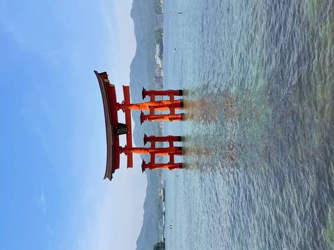       Vibrant red torii gate of Itsukushima Shrine standing in calm blue water with distant hills under a clear sky
  