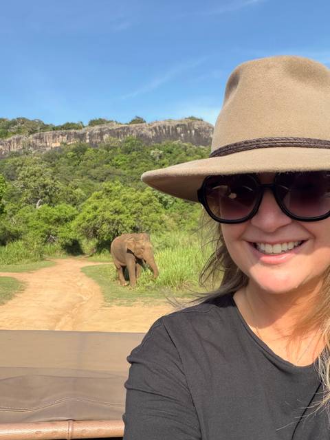       Close-up selfie of a smiling traveller in wide-brimmed hat with a wild elephant grazing on a dirt track behind.
  