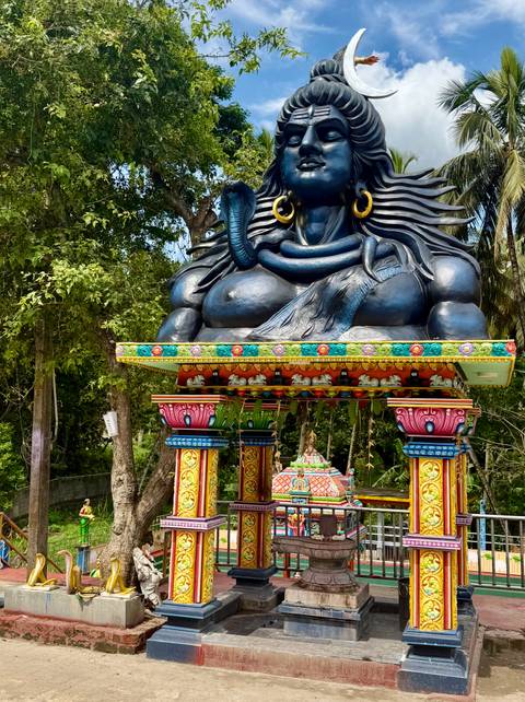       Vibrant multicoloured shrine featuring a large seated Shiva figure surrounded by jungle greenery.
  