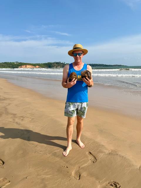       Traveller stands on an empty tropical beach holding freshly fallen coconuts with surf rolling behind.
  