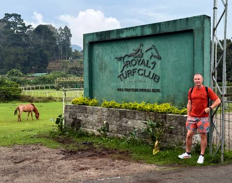       Visitor stands beside Royal Turf Club sign while a horse grazes on the lush track behind.
  