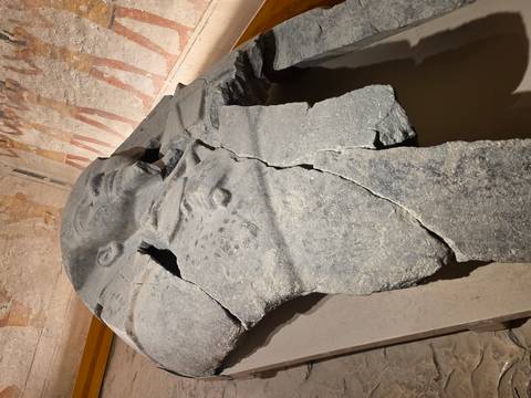       A shattered stone sarcophagus rests in a museum display against walls of ancient art.
  