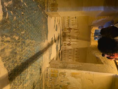       Tourists descend a decorated corridor inside an ancient Egyptian tomb with vivid blue and gold painted ceiling and wall reliefs.
  