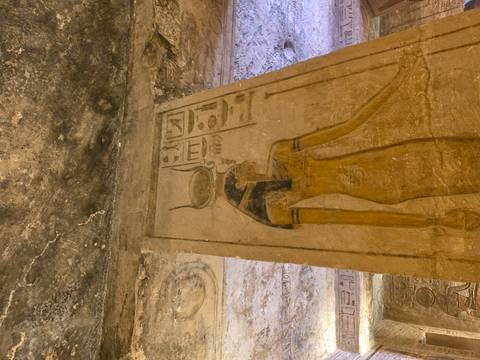       Painted relief of Egyptian goddess on column inside a tomb with yellow lighting.
  