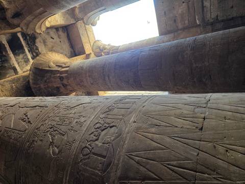      Tall decorated sandstone columns with hieroglyphs inside a dim temple hall.
  