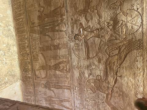       Temple wall relief showing chariot-riding pharaoh aiming bow among hieroglyphic panels.
  