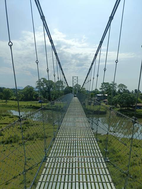       Metal suspension bridge stretching over a reflective river with verdant countryside beyond
  