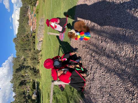       Women in traditional Andean dress sit beside a baby alpaca wearing a rainbow scarf
  