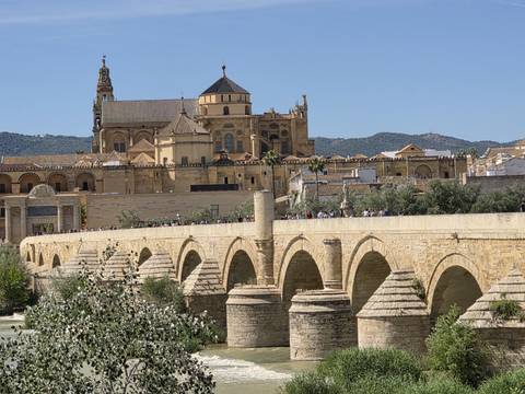       Stone Roman bridge crossing a river with the historic Mosque-Cathedral of Córdoba and mountains in the background.
  