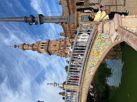       Ornate tiled bridge over a reflective canal with the towering Plaza de España in Seville under streaky blue skies.
  