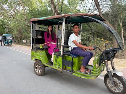      Passenger rides in a green e-rickshaw along a tree-lined Indian roadway.
  