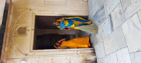       Two women in vibrant saris pause in palace doorway, one holding a sweeping broom
  