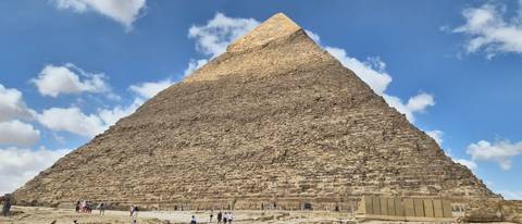       The Great Pyramid of Giza towering over visitors under a blue sky with clouds
  