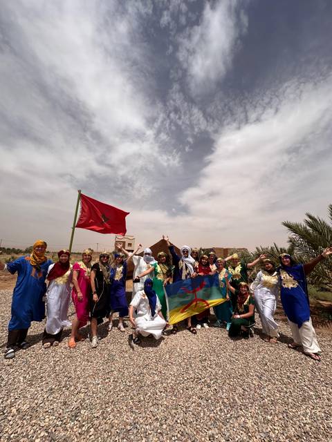       Large tour group in colourful traditional dress posing with Moroccan flags in an arid village setting
  