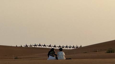       Two people sitting on dune watching long caravan of camels crossing desert ridge at dusk.
  