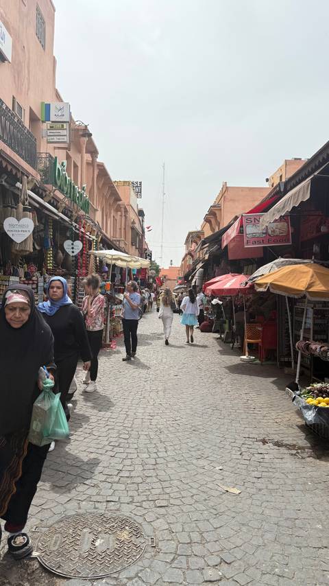       Bustling narrow souk lined with colorful stalls, umbrellas and shoppers in Marrakesh.
  