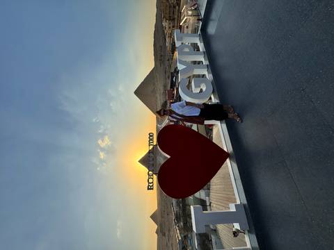       Traveler stands by a large 'I ❤️ EGYPT' sign on a rooftop with pyramids silhouetted at sunset.
  