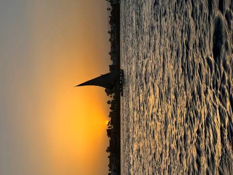      Traditional sailboat on rippling river water at golden sunset with urban skyline in silhouette.
  