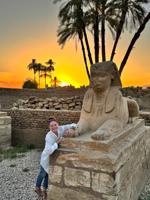       Smiling traveler leaning on a small sphinx statue amid ancient ruins with a vivid orange sunset and palm silhouettes
  