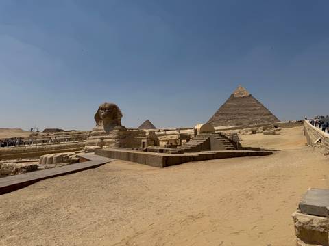       The Great Sphinx facing the Pyramid of Khafre under a clear blue sky with a wooden walkway leading through the sand
  