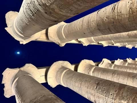       Illuminated stone columns of Luxor Temple reaching up to a deep blue night sky with a bright moon overhead
  