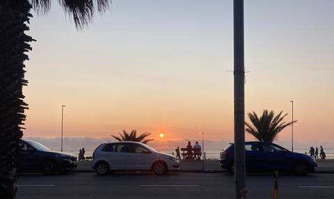       Sun setting over ocean boulevard with parked cars and silhouettes of palm trees.
  