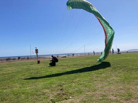       Paraglider canopy being laid out on a grassy seaside field while people stroll along the promenade
  