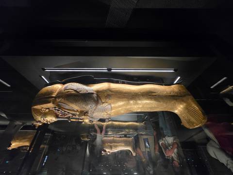       Golden pharaonic coffin encased in glass with onlookers in a darkened exhibit room.
  