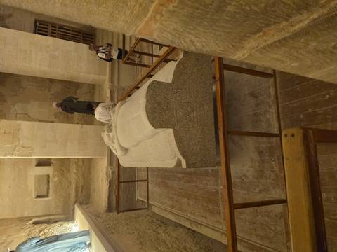       Stone granite sarcophagus displayed inside an ancient Egyptian tomb chamber with a few visitors observing.
  