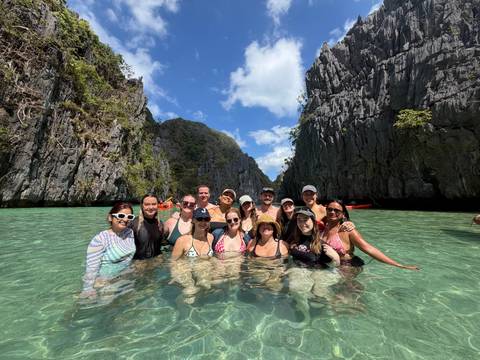       Group of swimmers pose in crystal-clear turquoise water flanked by towering limestone cliffs under a blue sky.
  