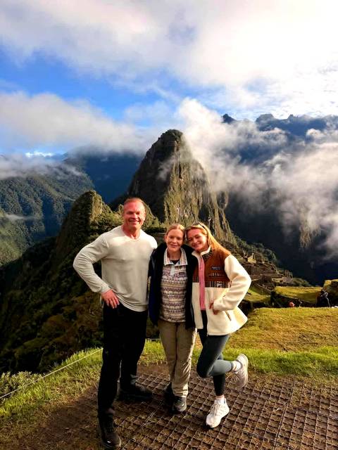       Three travellers smile in front of the mist-shrouded terraces and iconic peak of Machu Picchu.
  