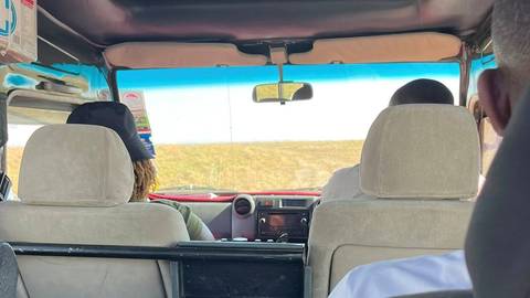       View from the rear seat inside a safari jeep looking through the windscreen toward open savannah.
  