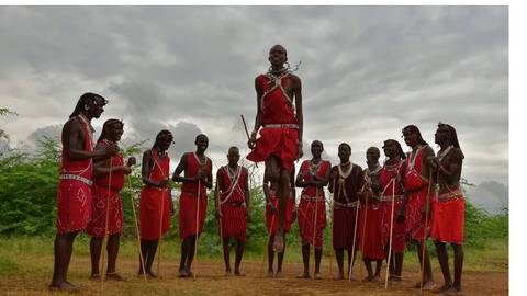       Maasai men dressed in red shukas performing a traditional jumping dance on dirt ground under cloudy skies.
  